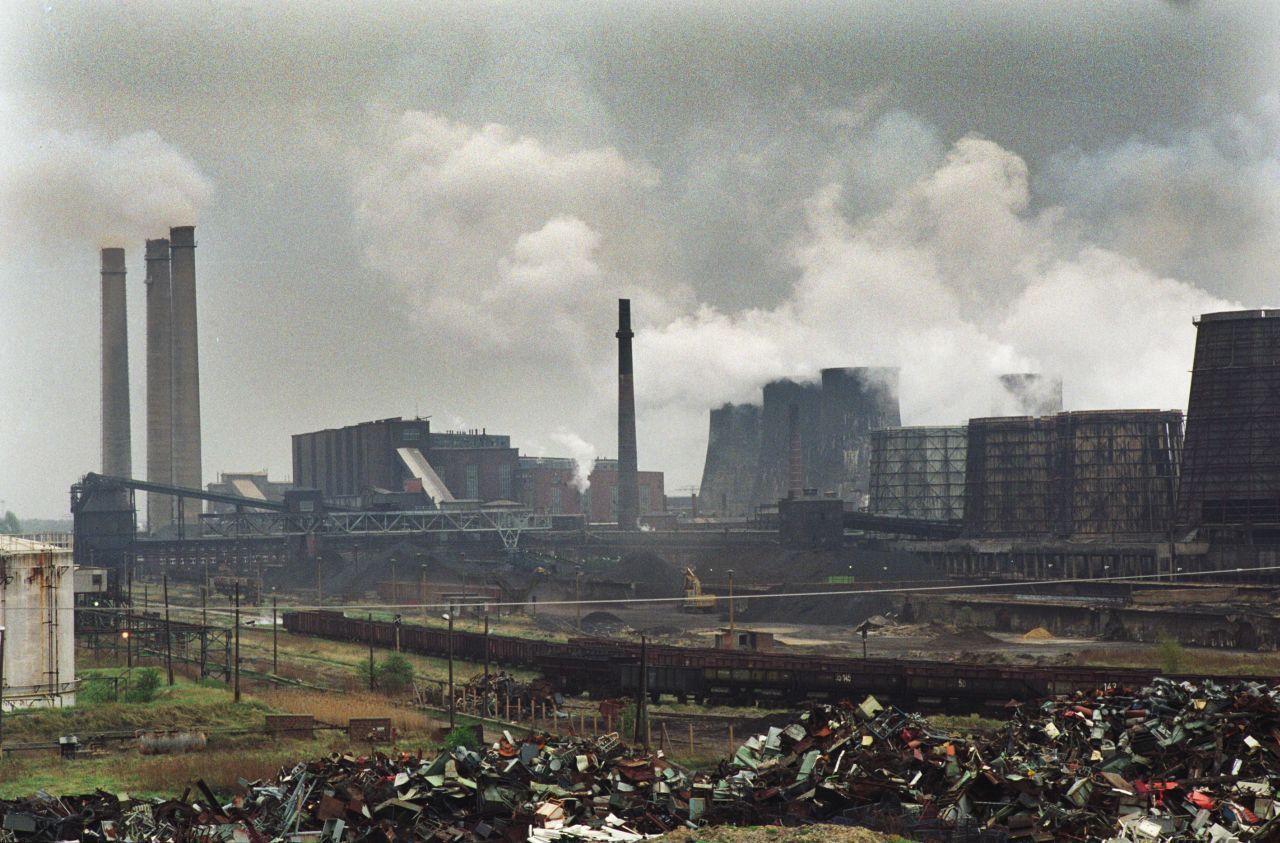 Farbfotografie des Braunkohlekraftwerks in Espenhain in Sachsen. Im Hintergrund dichte Abgaswolken über den Kraftwerktürmen, im Vordergrund hohe Müll- und Schuttberge.